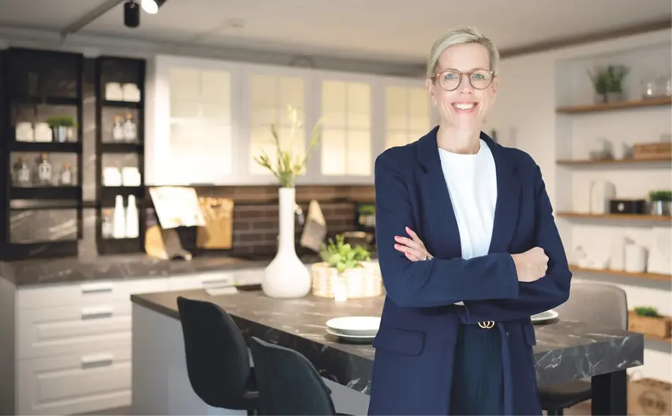 Frau mit Brille und verschränkten Armen steht vor Kücheninsel mit Stühlen, im Hintergrund moderne Einbauküche mit Regalen und Beleuchtung